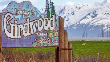 Uma placa de madeira com a inscrição 'Welcome to Chindwood Alaska', ao fundo montanhas e um campo gramado.