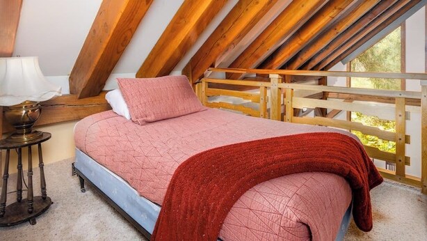 Un dormitorio con techo inclinado, una cama con una manta roja, una mesita de noche con una lámpara y vistas a la vegetación exterior.