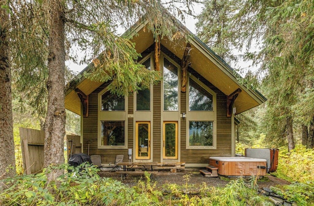 A cabin with a sloped roof, large windows, and a hot tub in a forest setting.