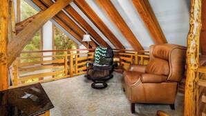 Una habitación con techo inclinado, vigas de madera, un sillón de cuero, una pequeña mesa y vistas amplias a través de grandes ventanas.