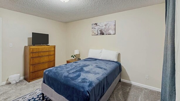 A bedroom with a bed, a dresser, a television on top, a nightstand with a lamp, and a painting on the wall. - Practical Anchorage Apartment Built for Families (Anchorage)