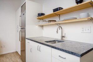 A modern kitchen with white cabinets, granite countertops, a stainless steel refrigerator, and wooden shelves above the sink.