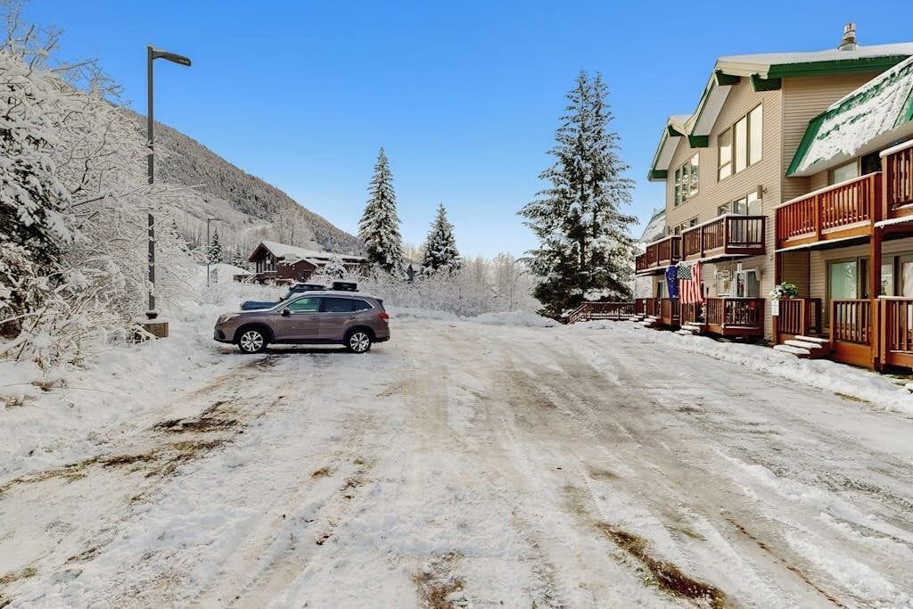 A snow-covered road with a car parked on the side, buildings with balconies, and snow-covered trees.