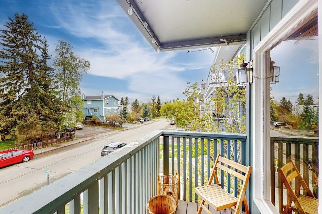 A balcony with a view of a street and houses, wooden chairs, a wicker basket, and a metal railing.