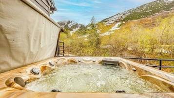 A hot tub with a wooden exterior, surrounded by a natural landscape with trees and a hill in the background.