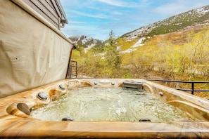 A hot tub with a wooden exterior, surrounded by a natural landscape with trees and a hill in the background. - Three-floor Slopeside Condo w/ Direct Mountain Views (Girdwood)