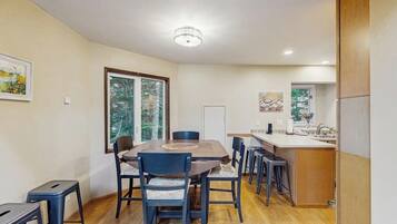A dining area with a wooden table and chairs, a kitchen with a sink and countertop, and a window with a view of trees.