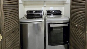 A laundry room with two stainless steel appliances, one dryer and one washer, enclosed by wooden louvered doors. - Lower Level of the Monarch Estate for Smaller Groups (Girdwood)