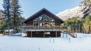 A wooden cabin with a balcony, surrounded by snow and trees, with a mountain in the background. - Lower Level of the Monarch Estate for Smaller Groups (Girdwood)
