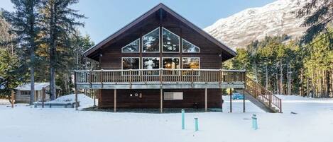 A wooden cabin with a balcony, surrounded by snow and trees, with a mountain in the background.