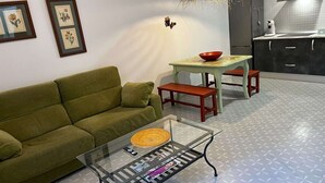 A living room with a green sofa, a glass coffee table with magazines, a dining area with a table and chairs, and a kitchen with a refrigerator and cabinets.