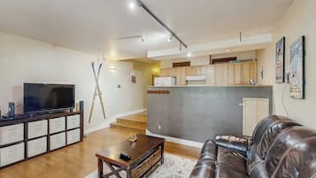 A living room with a leather couch, a TV on a stand, and a kitchen area in the background.