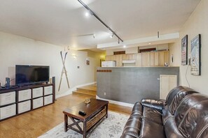 Una sala de estar con un sofá de cuero, un televisor sobre un mueble bajo, y una zona de cocina al fondo.