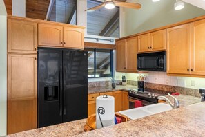 A kitchen with wooden cabinets, a black refrigerator, a microwave, granite countertops, and a ceiling fan.