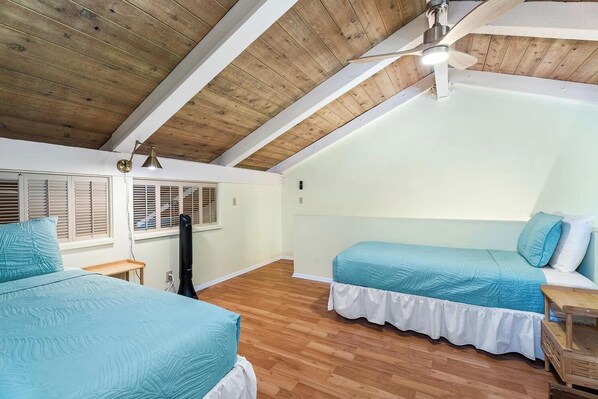 A bedroom with two beds, wooden flooring, a ceiling fan, and a window with shutters.