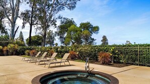 Una vasca idromassaggio circondata da poltroncine relax, con area piscina e una recinzione sullo sfondo adornata da piante.