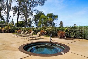 A hot tub surrounded by lounge chairs, with a poolside area and a fence with greenery in the background.