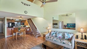 Un'ampia zona living con pavimento in legno, un divano con motivi tropicali, un tavolino da caffè e un'area pranzo con sedie in legno, con una cucina sullo sfondo.