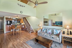 A spacious living area with a wooden floor, a sofa with tropical prints, a coffee table, and a dining area with wooden chairs and a kitchen in the background.