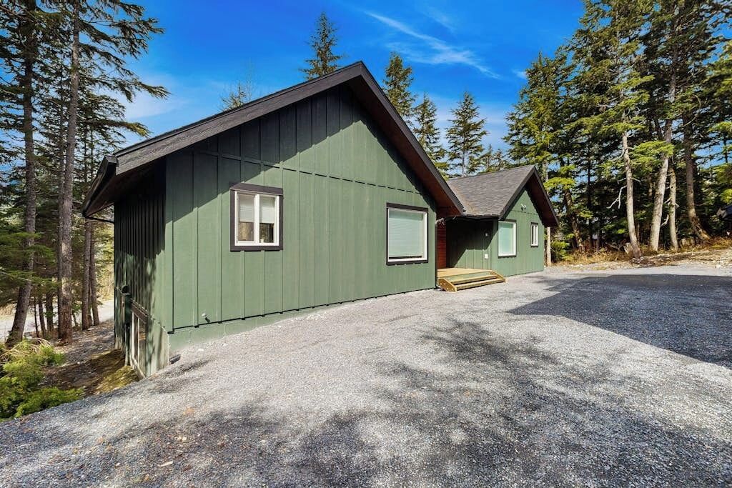 A two-story cabin with a gravel driveway, surrounded by tall trees.