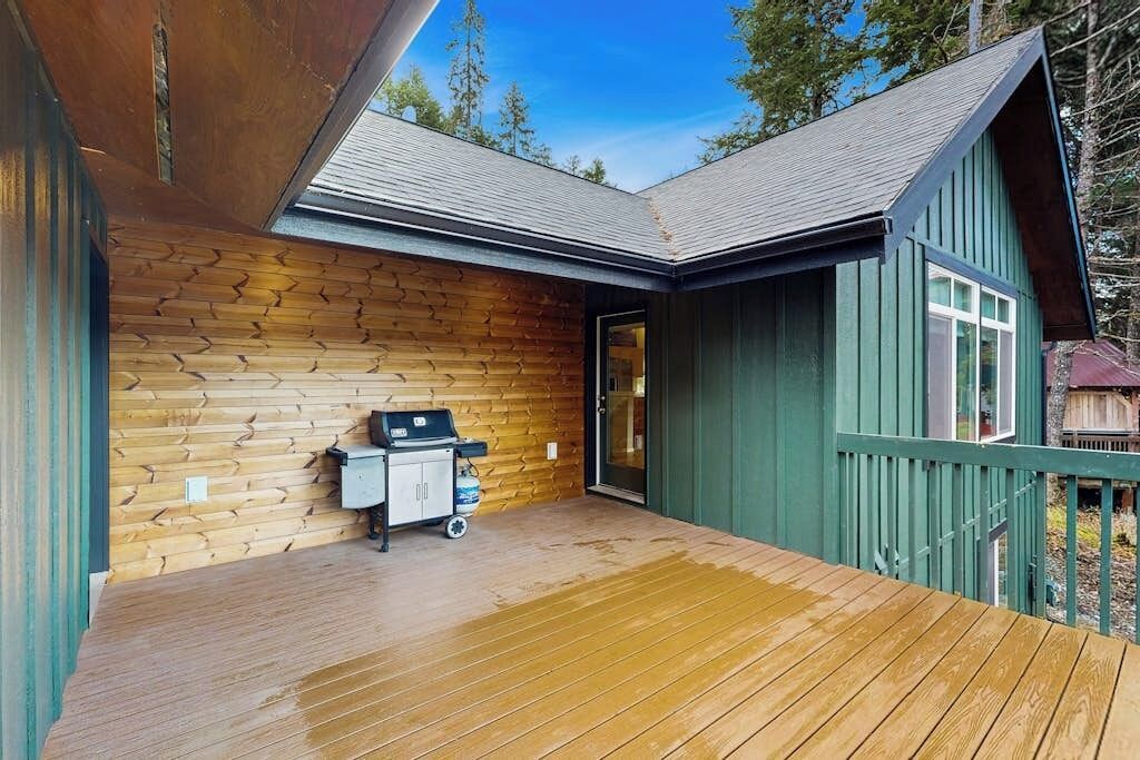 A wooden deck with a grill and a cart, a door leading to a covered area, and a green building with a sloped roof.
