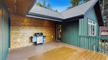 A wooden deck with a grill and a cart, a door leading to a covered area, and a green building with a sloped roof.