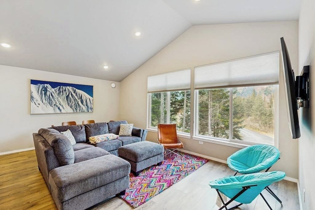 A modern living room with a sectional sofa, a colorful rug, a large window with a view, and a mounted TV.