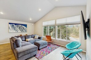 A modern living room with a sectional sofa, a colorful rug, a large window with a view, and a mounted TV.