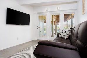 A modern living room with a flat-screen TV mounted on the wall, a brown leather sofa with a patterned pillow, and a glass door leading to an outdoor area.