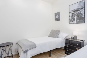A minimalist bedroom with a single bed, a bedside table, a small desk, and a wall-mounted picture.
