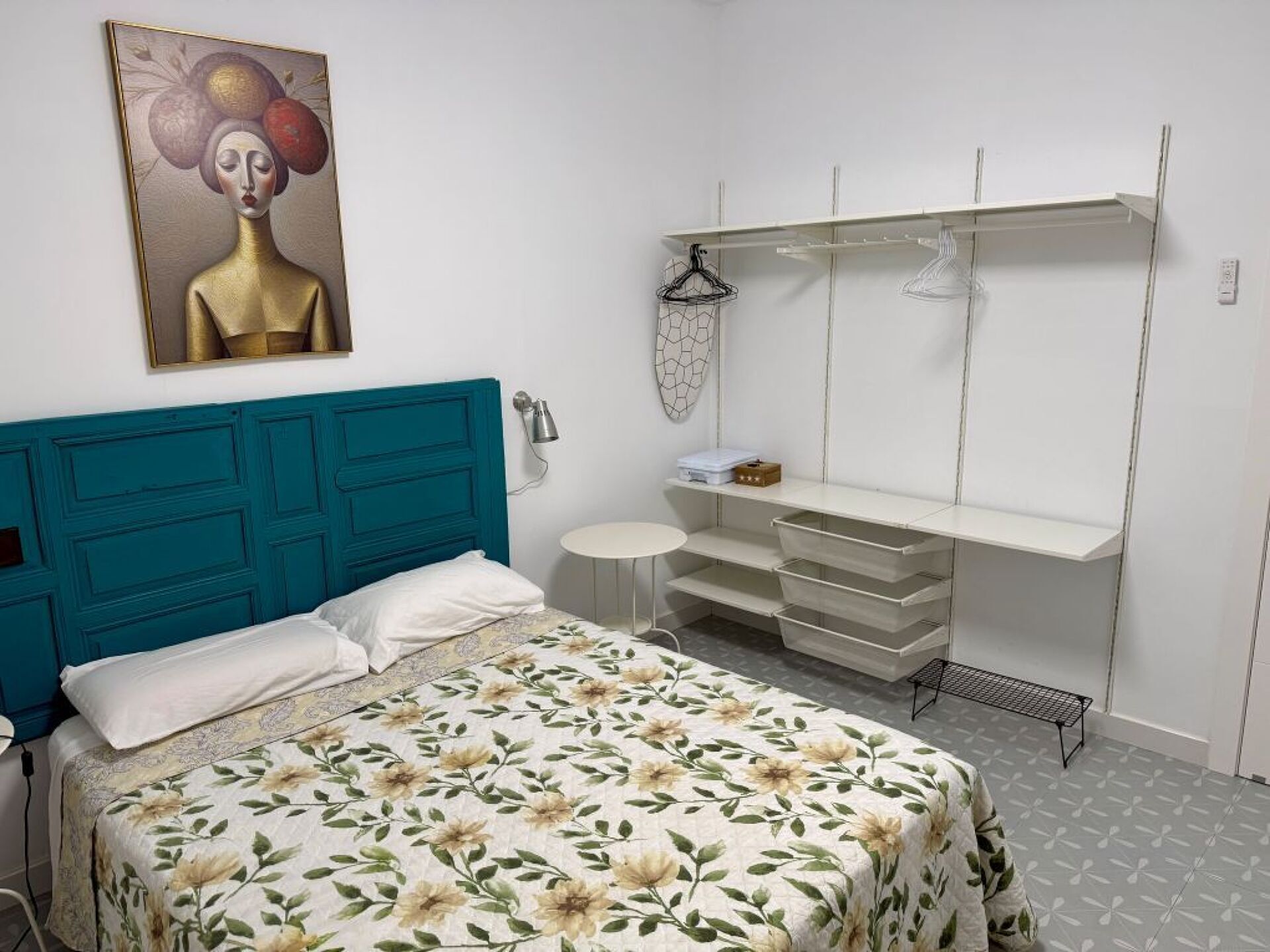 A bedroom with a teal headboard, a bed with floral bedding, a white dresser with drawers, and a wall-mounted mirror.