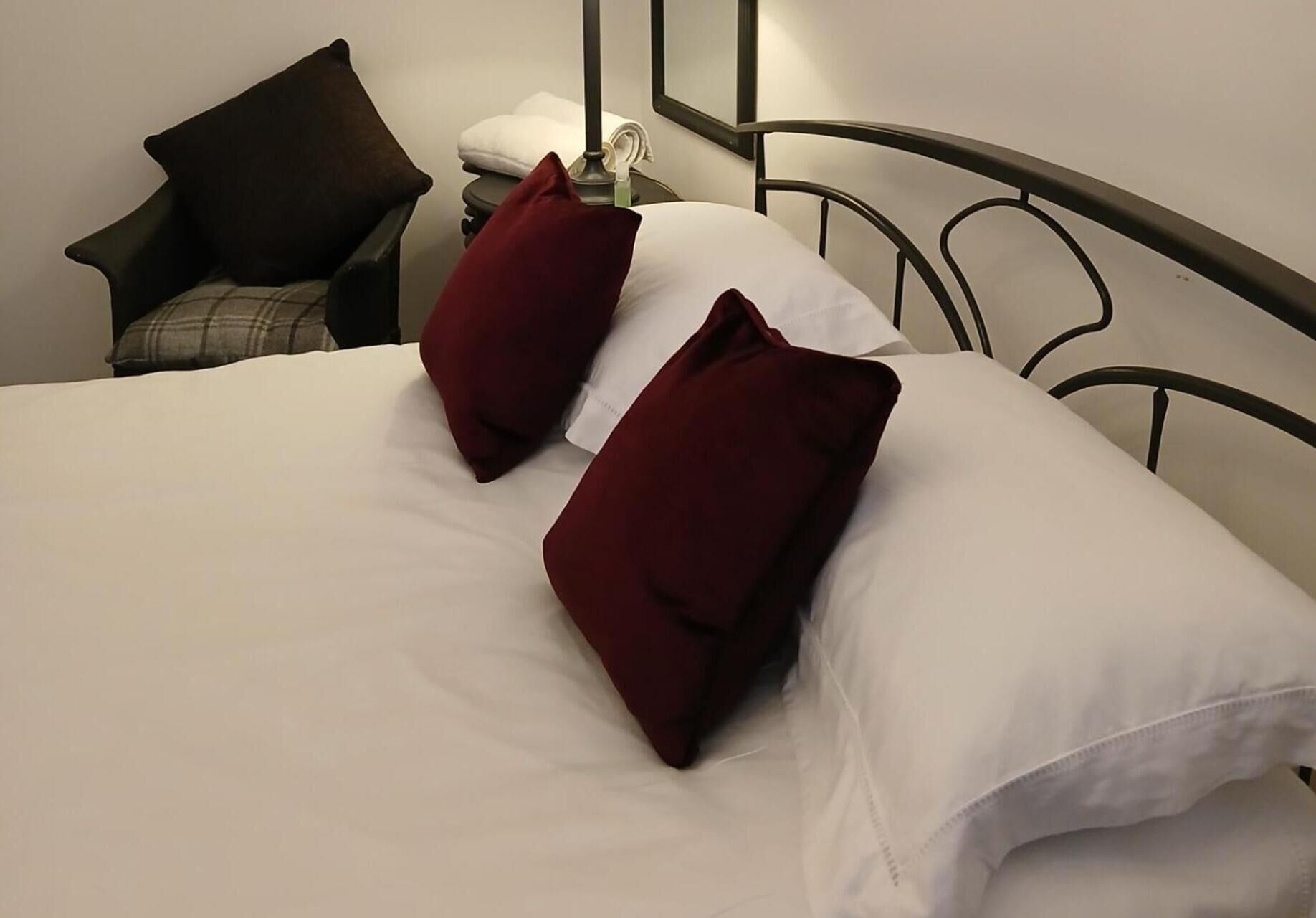 A bed with white bedding and two red pillows, a nightstand with a lamp, and a chair with a dark cushion.