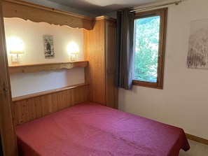 A room with a wooden headboard, a bed with a red bedspread, a window with a view of trees, and two wall-mounted lamps. - Apartment with parking - 6 guests (Valloire)