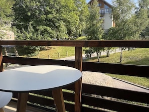 Een houten terras met een ronde tafel en stoelen, met uitzicht op een grasveld met bomen en een gebouw op de achtergrond.