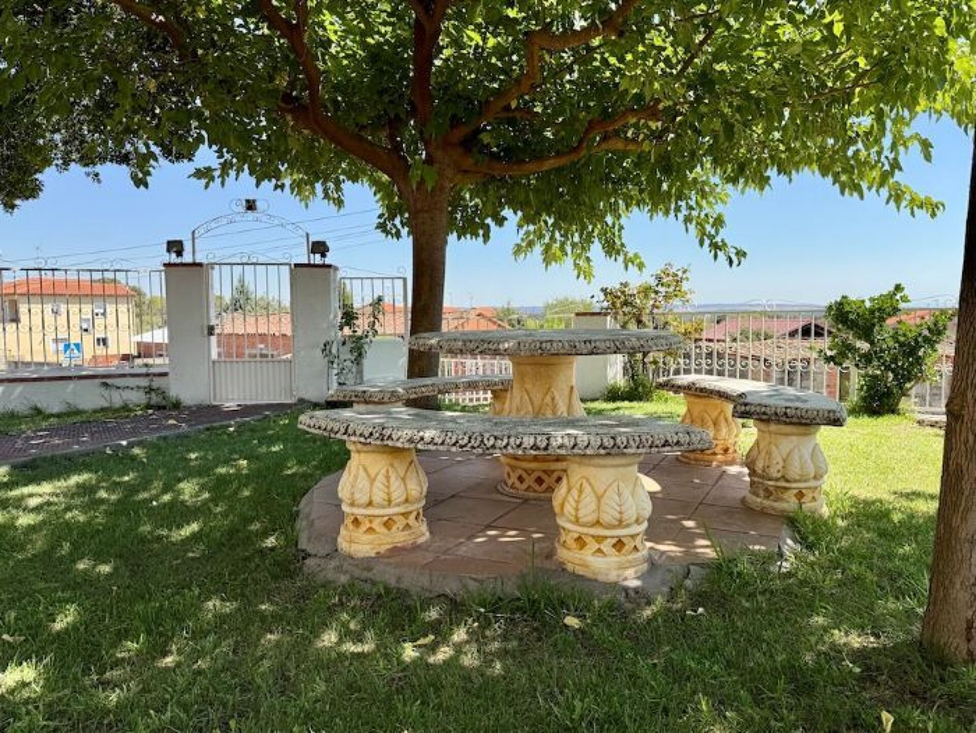Un jardin avec une table ronde en pierre, entourée de colonnes en pierre décorées, un arbre qui offre de l’ombre et une entrée fermée par un portail, visible au fond.