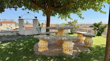 Un jardin avec une table ronde en pierre, entourée de colonnes en pierre décorées, un arbre qui offre de l’ombre et une entrée fermée par un portail, visible au fond.