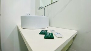 Un lavamanos blanco con un espejo encima, dos paquetes de champú verde y un paquete de jabón blanco en la encimera.