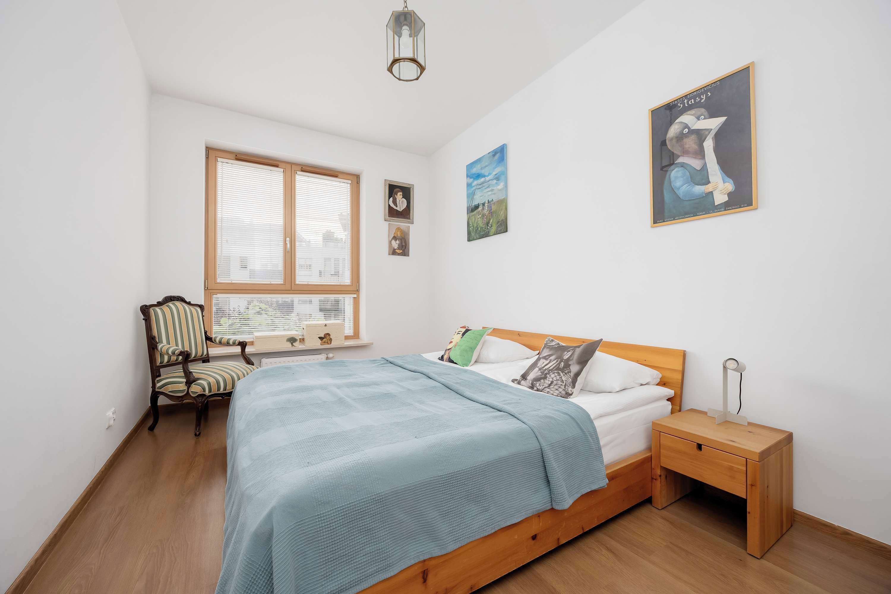 Une chambre à coucher comprenant un lit, une table de chevet, une commode, une chaise, une fenêtre et une œuvre d’art accrochée au mur.