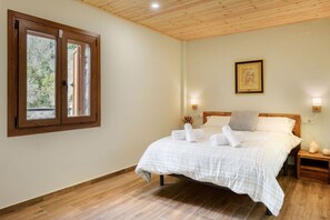 Een slaapkamer met een houten plafond, een bed met wit beddengoed, twee bedlampen en een raam met uitzicht op bomen.
