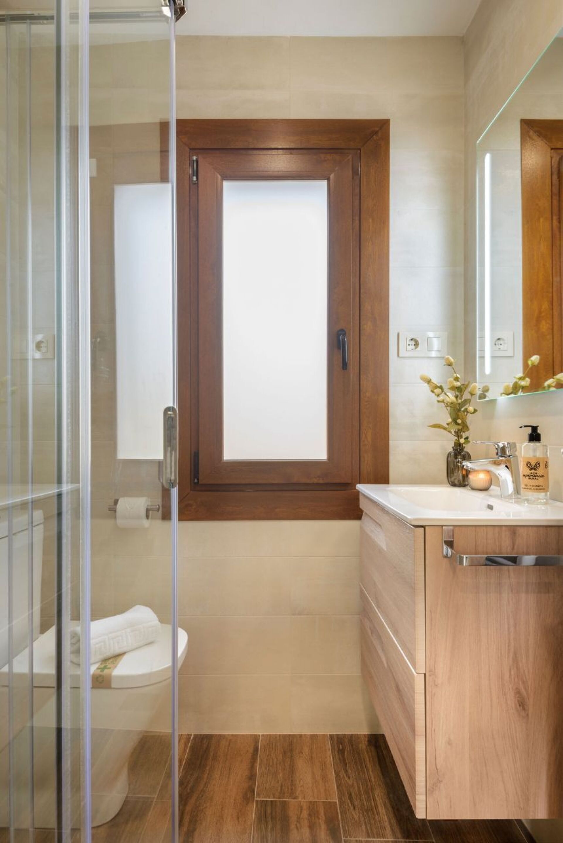 Une salle de bain avec une douche à parois de verre, une porte en bois, des toilettes blanches et un meuble-lavabo avec un miroir.