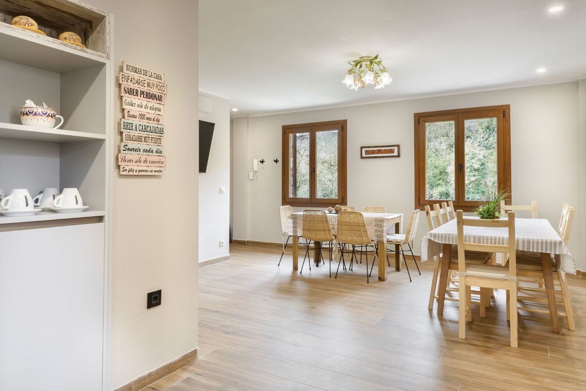 Un coin repas avec une table dressée pour quatre personnes, entourée de chaises. On y trouve une étagère avec des tasses et un panneau avec du texte accroché au mur.