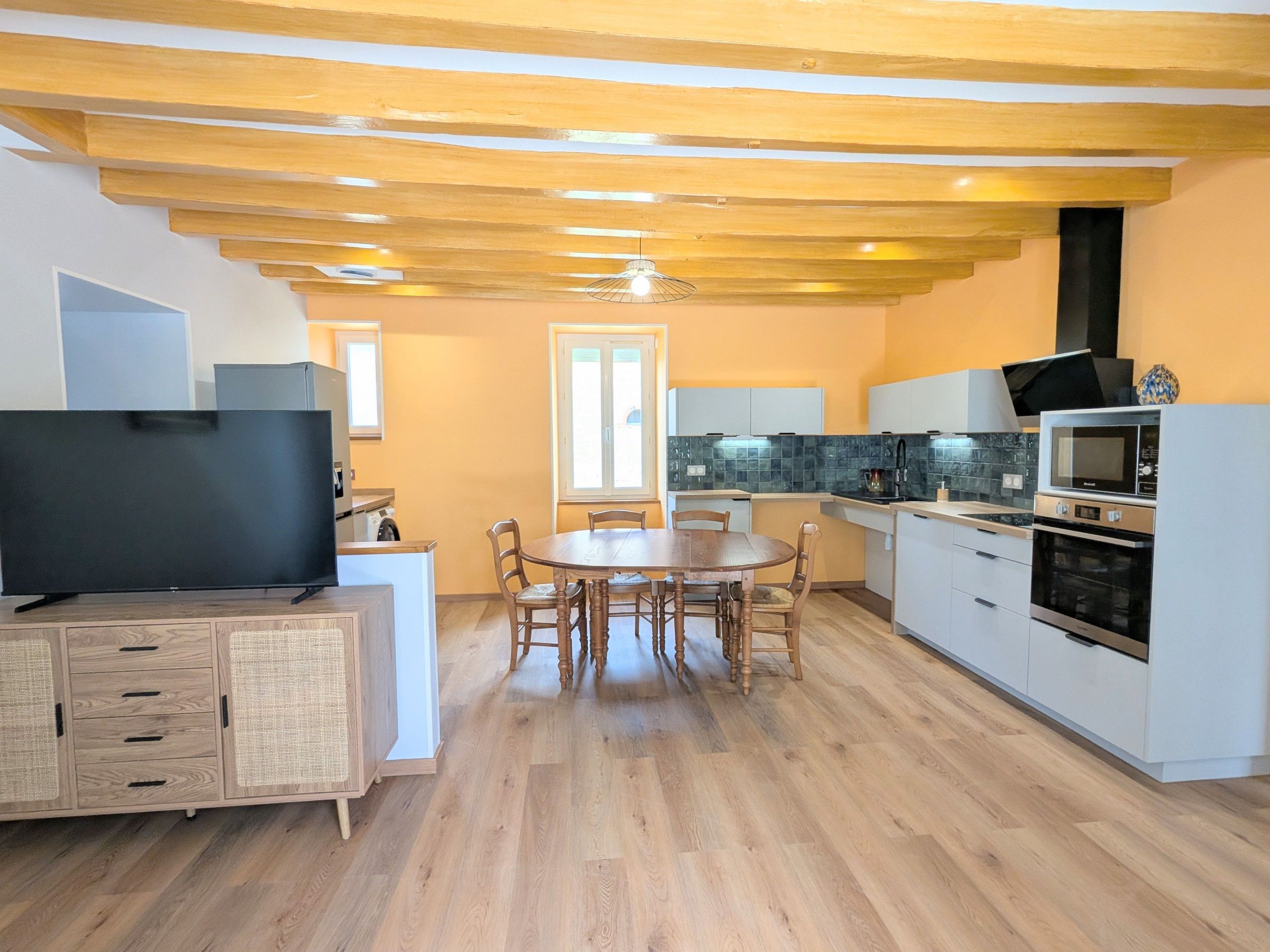Cozinha compacta com eletrodomésticos modernos, área de refeições com mesa e cadeiras de madeira, e televisão em um suporte.