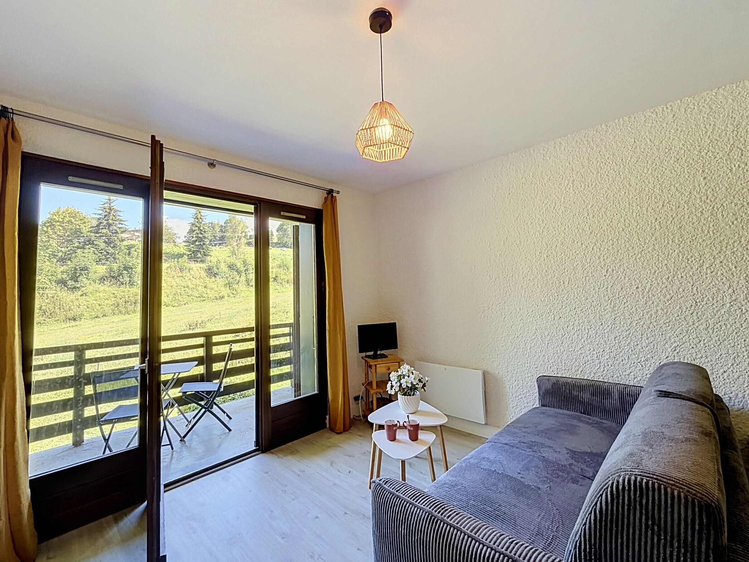 Una habitación con sofá, una mesita con un jarrón, televisor y vistas a un paisaje verde a través de amplias ventanas.