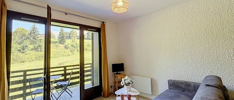 Una habitación con sofá, una mesita con un jarrón, televisor y vistas a un paisaje verde a través de amplias ventanas.