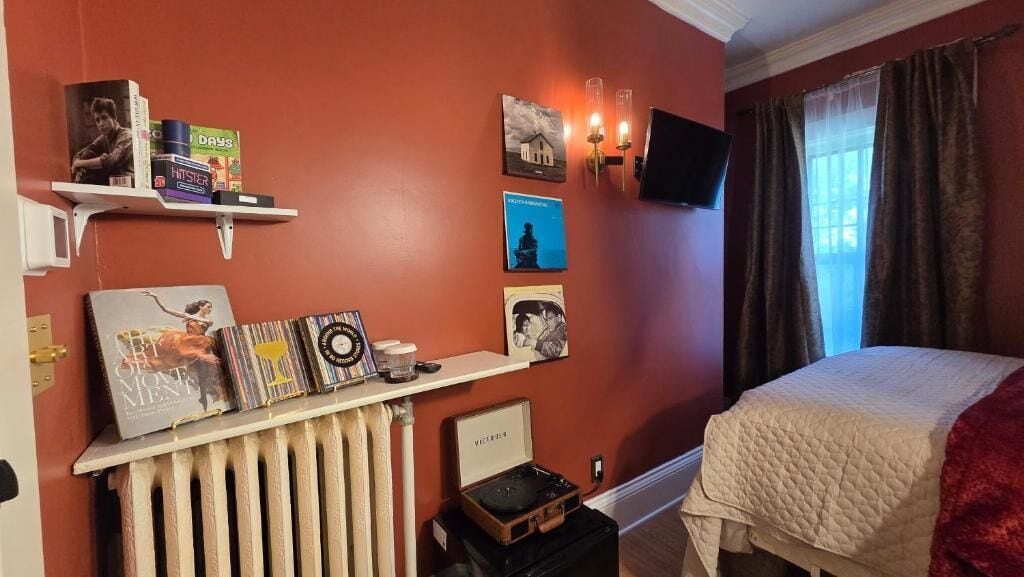 A bedroom with a bed, a radiator, a shelf with books and a record player, a wall-mounted TV, and a window with curtains.