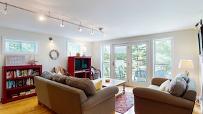 A spacious living room with a sofa set, a red entertainment unit, and a dining table. - Waterfront Retreat with Private Dock, Kayaks & Stunning Water Views (Phippsburg)