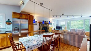 A modern kitchen with a dining area, a stone fireplace, and a large window overlooking greenery. - Waterfront Retreat with Private Dock, Kayaks & Stunning Water Views (Phippsburg)