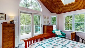 A bedroom with a wooden bed, a green chair, and a view of the outdoors through large windows. - Waterfront Retreat with Private Dock, Kayaks & Stunning Water Views (Phippsburg)