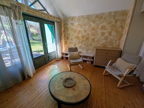 Ein Zimmer mit einem runden Tisch, zwei Stühlen, einem Holzschrank und einem großen Fenster mit Vorhängen.