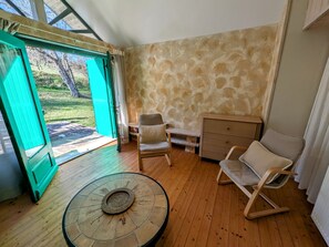 Una habitación con piso de madera, una mesa redonda con un centro de mesa tejido, dos sillas, un armario de madera y una vista a través de una puerta abierta hacia un espacio con césped.